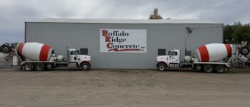 Two white and red concrete mixer trucks are parked in front of a large gray industrial building with a sign that reads "Buffalo Ridge Concrete Inc" in Watertown, SD. The ground is covered in packed dirt, and the sky is overcast.