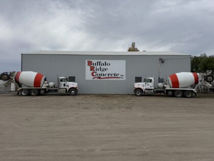 Two white and red concrete mixer trucks are parked in front of a large gray industrial building with a sign that reads "Buffalo Ridge Concrete Inc" in Watertown, SD. The ground is covered in packed dirt, and the sky is overcast.